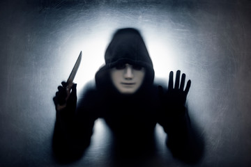 Man in a hood and white mask with knife behind a dusty scratched glass