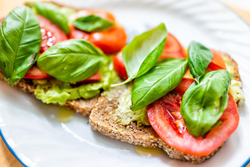 Closeup of two slices pieces of whole wheat sprouted toasted grain bread on plate with red tomato slices green basil leaves and avocado bruschetta on plate sesame seeds