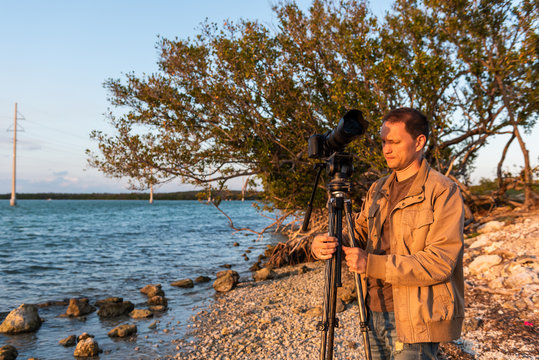 Sunset in Florida Keys rocky beach with young man photographer standing setting up tripod taking picture looking at coastline coast sunlight