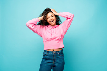 cheerful curly african american woman touching hair isolated on blue