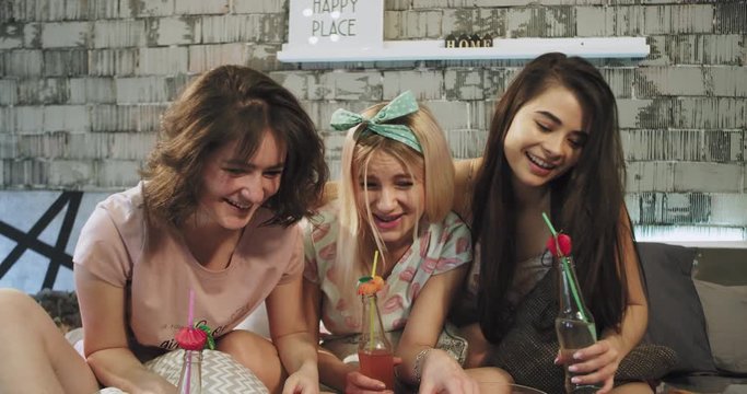 Very charismatic with a large smile teenager girls closeup on the bed have a sleepover , spending a good time together drinking some yummy drinks from the bottle and chatting.