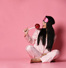 Beautiful young brunette woman in pajama hold huge sweet lollypop on pink background 