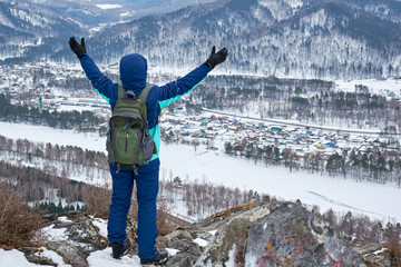 Tourist girl with a backpack standing on top of a mountain in winter clothes. A hood on her head lifts her hands upwards rejoices the landscape that opens to her. hiking in the mountains in winter