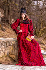 Obraz premium Art Photography. Mysterious Medieval Queen in Red Dress and Spiky Black Crown Posing With Ancient Book in Forest in Early Spring.