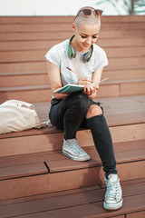 Smart young college student, urban woman, writing down some notes in her notebook, sitting outdoors on a beautiful sunny day