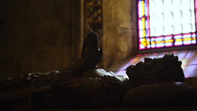 Crop view monument on stone coffin with remains of Marco Polo in cathedral. Sculpture in dark space illuminated colorful light from stained glass window. Lisbon, Portugal