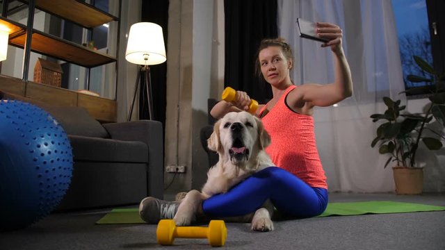 Attractive sporty female holding dumbbell taking selfie pictures with labrador pet on cellphone after productive workout at home. Joyful fit woman ready to post selfie shots on social media