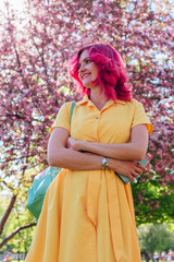 Beautiful and young woman with bright red hair and ref lips standing next to a blooming apple tree in a yellow dress.