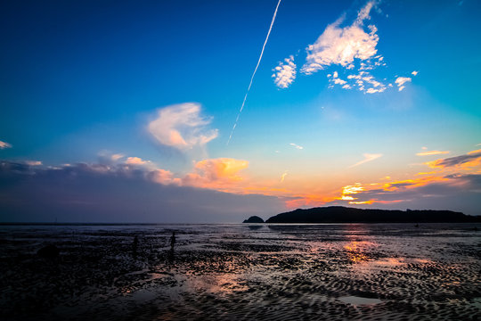 Far-away view of a muddy shore reflecting blue and orange gradient sky with dramatic sunset with airplane trail