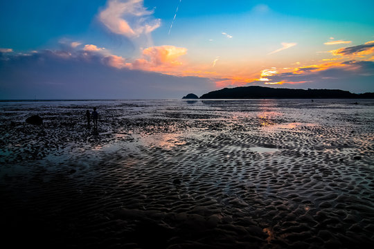 Far-away view of a muddy shore reflecting blue and orange gradient sky with dramatic sunset with airplane trail