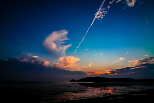 Far-away view of a muddy shore reflecting blue and orange gradient sky with dramatic sunset with airplane trail