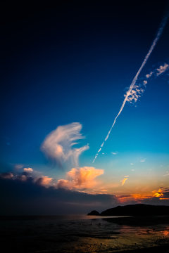 Far-away view of a muddy shore reflecting blue and orange gradient sky with dramatic sunset with airplane trail