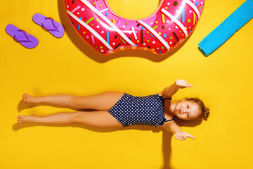 Top view of a little girl lying on the floor with slippers, a towel and an inflatable circle donut on the side. A child in a bathing suit is resting on a yellow background and shows his thumb.