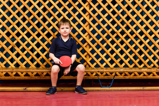 seven-year-old Caucasian boy in gray sports uniform sitting on a wooden bench and holding a racket for table tennis