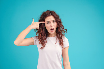 Attractive brunette girl with curly hair isolated over blue turquoise background shooting herself with finger