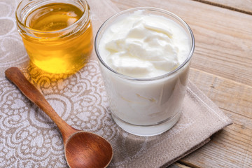 Transparent glass of  fat homemade yogurt with spoon and jar of honey stands on wooden table.
