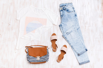 White t-shirt with print, light blue ripped boyfriend jeans, beige sandals, textile striped bag on white wooden background. Overhead view of woman's casual outfits. Flat lay, top view.