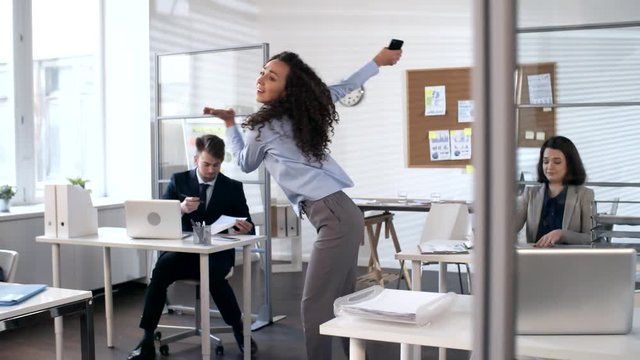 Young businesswoman talking on phone and then walking through the office, smiling and dancing in front of colleagues while celebrating good news