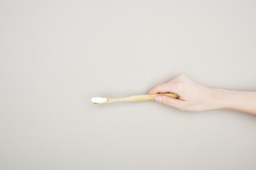 partial view of woman holding bamboo toothbrush with toothpaste on grey background