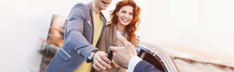 Fototapeta na wymiar panoramic shot of car dealer giving keys to cheerful man and woman in car showroom