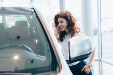 cheerful curly woman standing with hand in pocket near automobile