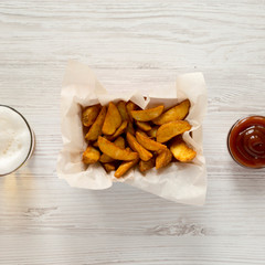 Fried potato wedges in paper box, barbecue sauce and glass of beer on a white wooden table. Flat lay. Overhead, top view, from above.