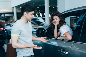 cheerful woman sitting in automobile near handsome man standing and gesturing in car showroom