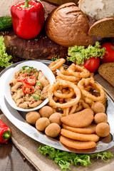 Assorted beer snacks: onion rings, cheese balls, chips etc on a iron tray on a dark wooden background among vegetables. Close-up. Space. 