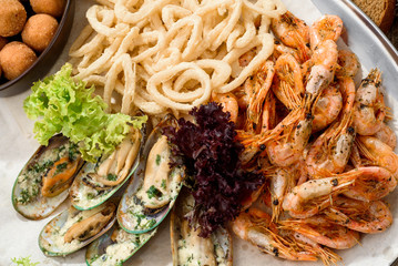 Assorted beer snacks: onion rings, mussels in a creamy sauce, fried shrimps and cheese balls on craft paper on a tray. Composition on a dark wooden background with fresh vegetables. Close-up. Space.