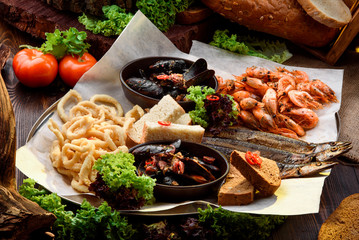 Assorted beer snacks: onion rings, smoked fish, mussels and shrimps on a tray on sackcloth among vegetables. Bread on a wooden background. Close-up. Space.