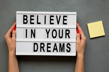 Female hands hold modern board with 'Believe in your dreams' words over gray background. From above, overhead, flat lay.