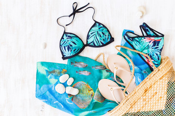 Blue and green swimsuit with tropical print, beige flip flops and straw wicker beach bag on white wooden background. Overhead view of woman's swimwear and beach accessories. Flat lay, top view.