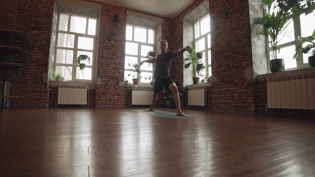 Man practice warrior yoga pose in studio with brick wall. Sporty man doing yoga indoors with wooden floor and big windows with copyspace. Wide angle