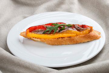 Bruschetta with anchovies and grilled pepper on a white plate, on a textile background