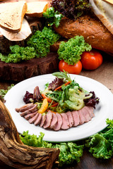 Duck slices with lettuce and orange slices on a white plate on a dark wooden table among vegetables. Close-up.