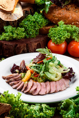Duck slices with lettuce and orange slices on a white plate on a dark wooden table among vegetables. Close-up.