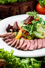 Duck slices with lettuce and orange slices on a white plate on a dark wooden table among vegetables. Close-up.