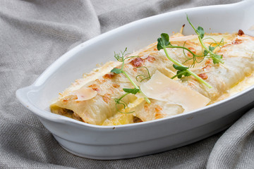 Cannelloni with minced meat, baked in a white ceramic plate. On textile background