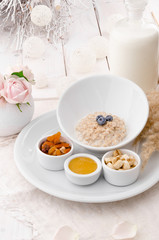 Oatmeal with blueberries in a beautiful white plate with honey, berries and a banana on a light delicate background, decorated with live roses. Beautiful healthy breakfast. Close-up. Space