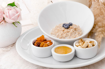 Oatmeal with blueberries in a beautiful white plate with honey, berries and a banana on a light delicate background, decorated with live roses. Beautiful healthy breakfast. Close-up. Space