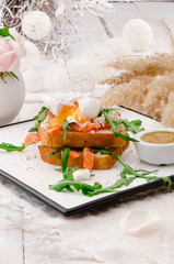 Breakfast: white bread toasts with salted salmon, poached egg and arugula, served with sots on a white plate. Beautiful light composition with fresh roses. Close up.  Space