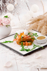 Breakfast: white bread toasts with salted salmon, poached egg and arugula, served with sots on a white plate. Beautiful light composition with fresh roses. Close up.  Space