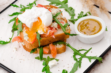 Breakfast: white bread toasts with salted salmon, poached egg and arugula, served with sots on a white plate. Beautiful light composition with fresh roses. Close up.  Space