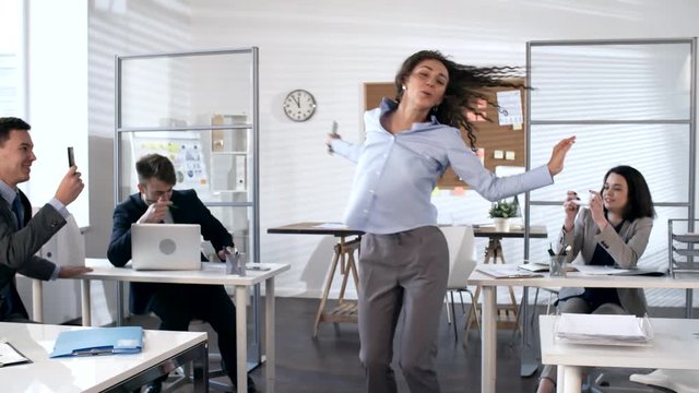Beautiful young businesswoman happily smiling and dancing in the center of office while cheerful colleagues taking pictures of her with smartphones