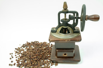 Vintage rusty, wooden black pepper grinder or mill with a mechanical handle  gear and scattered whole dried black pepper on white background.