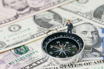 Miniature businessman in suit standing and thinking about future on compass and pile of US dollar money banknote, investment, financial crisis or economic direction concept