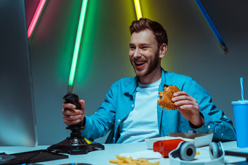 handsome and smiling man holding tasty burger and playing video game with joystick