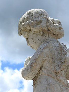 Scene in a cemetery: close-up of an old stone statue of a little angel. The statue is damaged by the passage of time. Cloudy sky on a sunny day. Faith and religiosity.