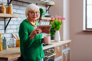 Aged woman looking at picture frame and enjoying morning coffee