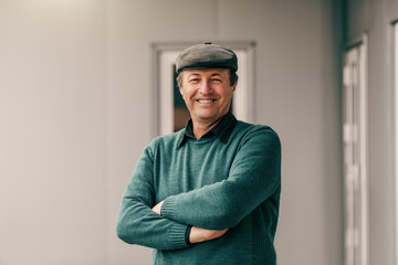 Smiling male senior with cap on head and with arms crossed standing in front of house at spring.